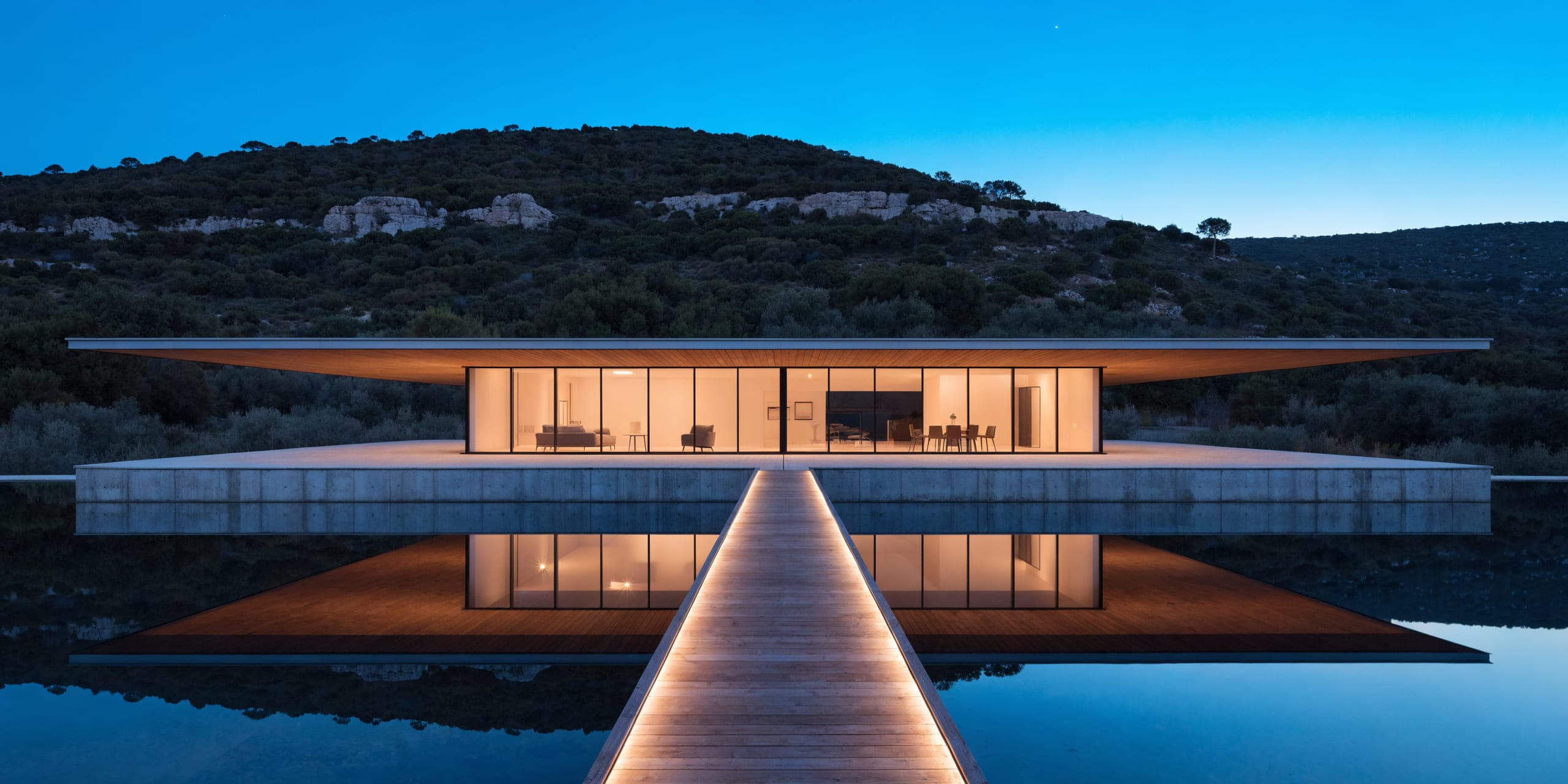 A villa at dusk mirrored in a reflecting pool, lit from within